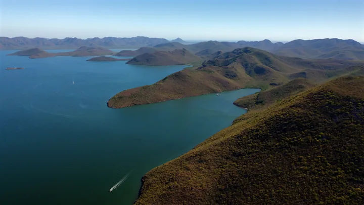 Vista aérea de una bahía amplia rodeada de cerros bajos y penínsulas, con una lancha dejando estela sobre el agua