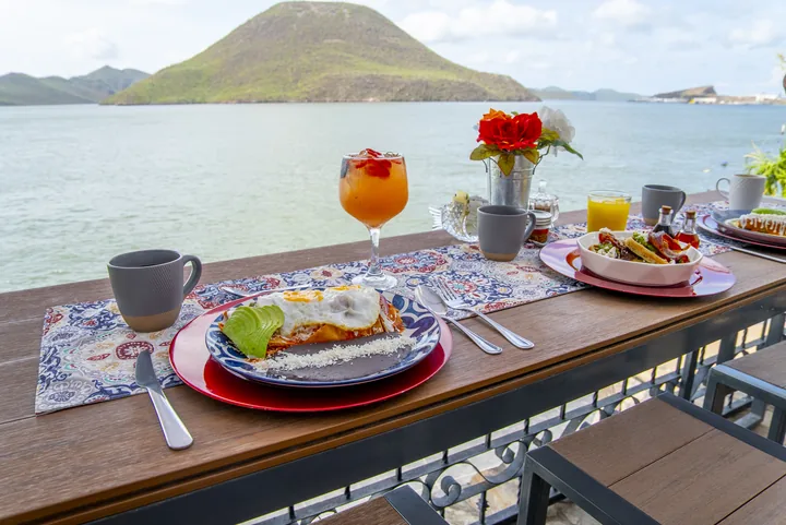 Desayuno servido en la terraza de un restaurante frente a una bahía, con chilaquiles, molletes y agua fresca, y un cerro verde al fondo