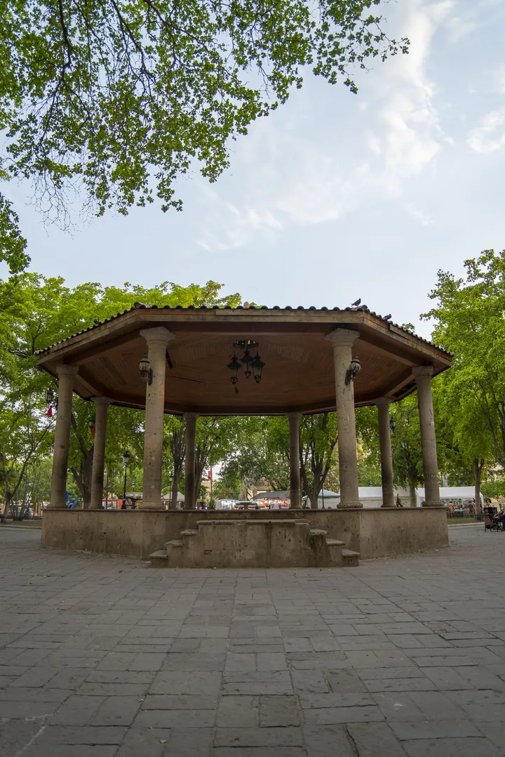 Kiosco octagonal de columnas de cantera y techo de teja en una plaza pública arbolada, con piso de adoquín
