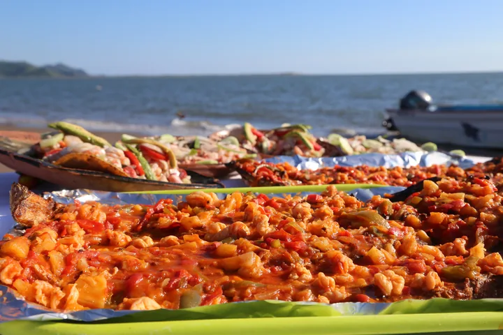 Bandejas de mariscos preparados sobre una mesa en la playa, con una lancha y el mar al fondo