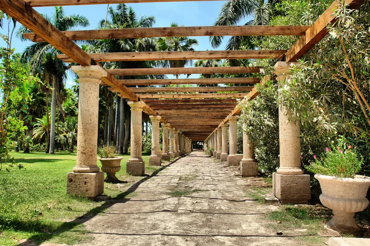 Pérgola de columnas de cantera y vigas de madera sobre un andador de piedra, entre palmeras y jardines de un parque