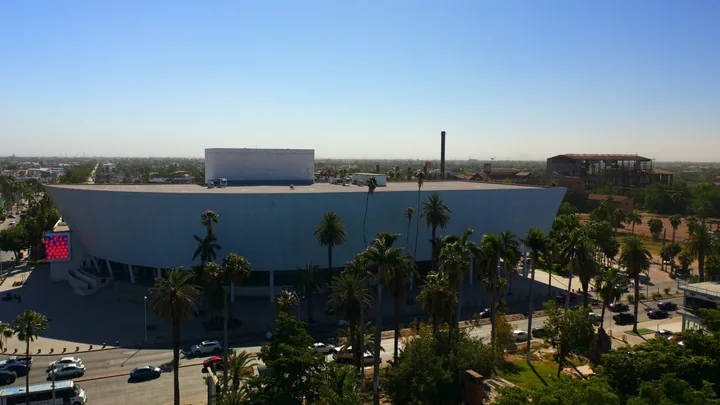 Vista aérea de un edificio de forma curva rodeado de palmeras, con la chimenea de ladrillo del antiguo ingenio al fondo