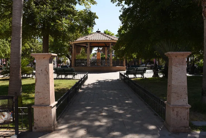 Andador de la plazuela hacia un kiosco de teja rodeado de árboles y bancas