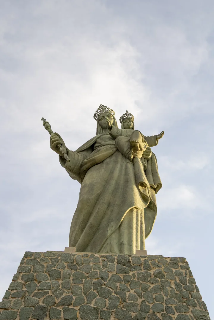 Escultura de la Virgen con el Niño, coronados y sobre una base de piedra, vista a contraluz contra el cielo